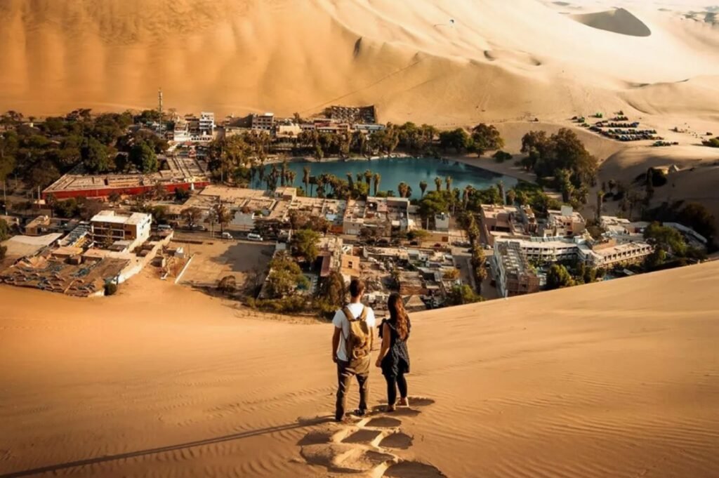 Dos viajeros frente al oasis de Huacachina rodeado de dunas doradas en Ica